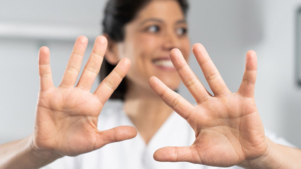 Egy fehér laborköpenyt viselő ember a kamerába néz Mindkét keze kinyújtva, széttárt ujjakkal A személy arca szándékos elhomályosítva a személyazonossága védelméért. A háttérben elmosódott beltéri környezet egy falon lógó képpel és egy ágynak vagy kanapénak látszó bútordarabbal.