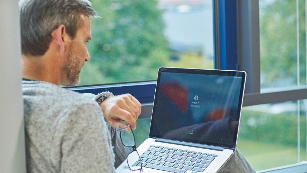 Egy ülő ember laptopot tart az ölében Bal kezével gépel, jobb kezében pedig szemüveget tart. A laptop képernyőjén egy elmosódott fejhallgató ikon látható a kattintson a meghallgatáshoz címkével A háttérben egy zöld, természetes táj látható egy széles ablakon keresztül.