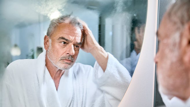 idős férfi fürdőköpenyben figyelmesen néz bele a ködös fürdőszobatükörbe