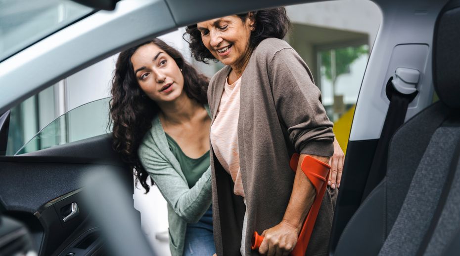 Egy mankót használó beteg, és egy másik személy, aki segít neki beszállni a járművébe.