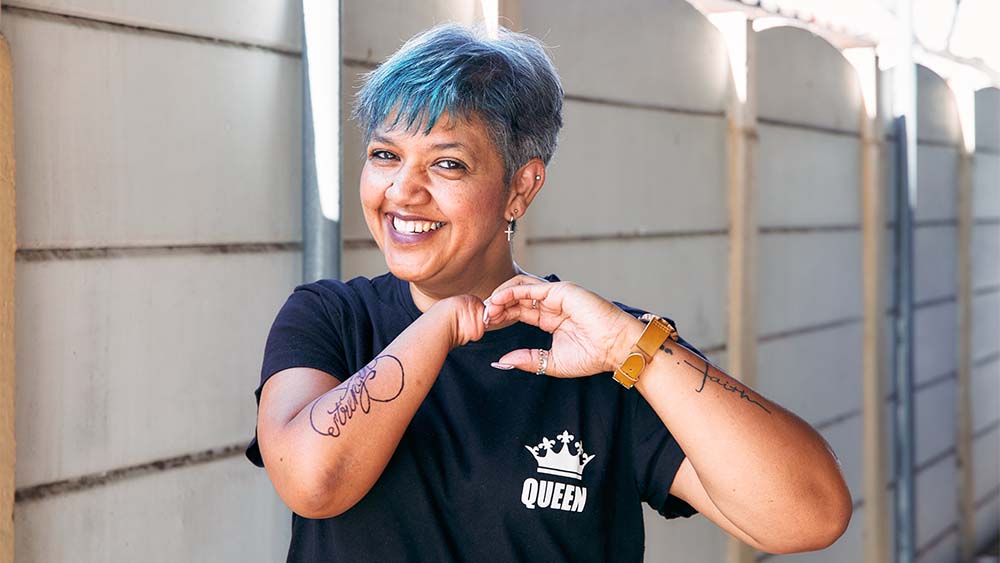 Egy nő jobb kéz nélkül, kék hajjal és tetoválással mosolyog, magabiztosságot és önkifejezést sugároz. 